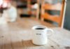 Background blurred of a wooden table. In the foreground is a white coffee cup that says, "Begin."