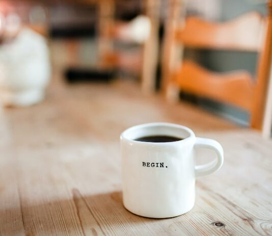 Background blurred of a wooden table. In the foreground is a white coffee cup that says, "Begin."
