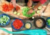 Curved wood-style cutting board and close up of hands preparing lettuce, tomatoes, red peppers, and other items in blue and black bowls of different sizes. Hands are light skimmed and one has a knife cutting lettuce and is wearing a watch. Photo Source: Maarten van den Heuvel, @mvdheuvel, via Unsplash.