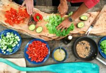 Curved wood-style cutting board and close up of hands preparing lettuce, tomatoes, red peppers, and other items in blue and black bowls of different sizes. Hands are light skimmed and one has a knife cutting lettuce and is wearing a watch. Photo Source: Maarten van den Heuvel, @mvdheuvel, via Unsplash.