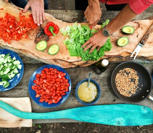 Curved wood-style cutting board and close up of hands preparing lettuce, tomatoes, red peppers, and other items in blue and black bowls of different sizes. Hands are light skimmed and one has a knife cutting lettuce and is wearing a watch. Photo Source: Maarten van den Heuvel, @mvdheuvel, via Unsplash.