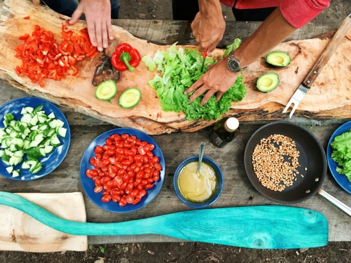 Curved wood-style cutting board and close up of hands preparing lettuce, tomatoes, red peppers, and other items in blue and black bowls of different sizes. Hands are light skimmed and one has a knife cutting lettuce and is wearing a watch. Photo Source: Maarten van den Heuvel, @mvdheuvel, via Unsplash.